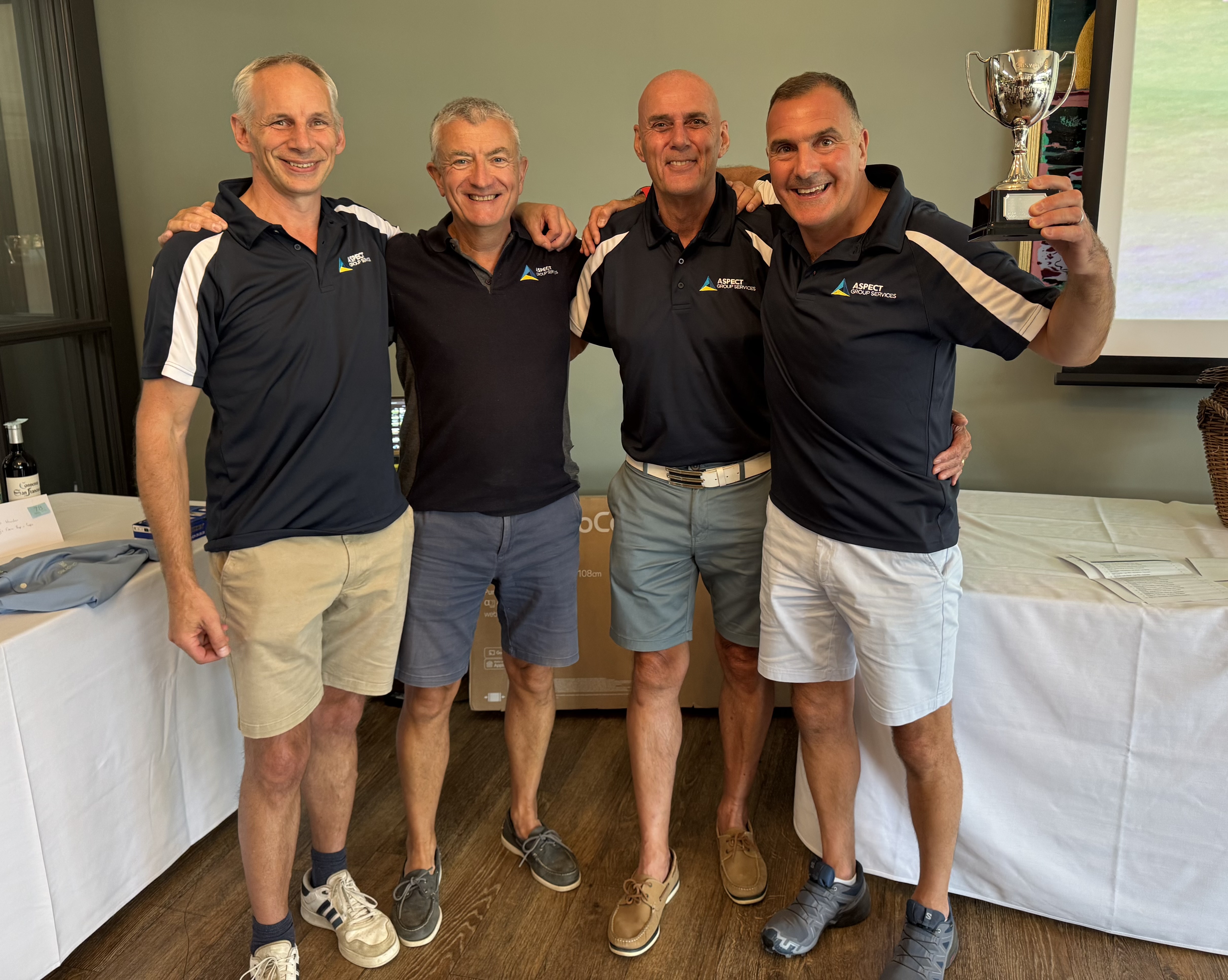 The 4-man team from Aspect Group Services stand side-by-side. All 4 men are in navy blue polo shirts and short, with the person on the right holding a trophy for the team's win